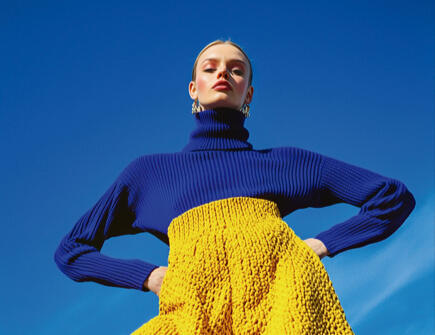 AI-generated fashion model posing against a bright blue sky, wearing a striking yellow and blue avant-garde outfit.